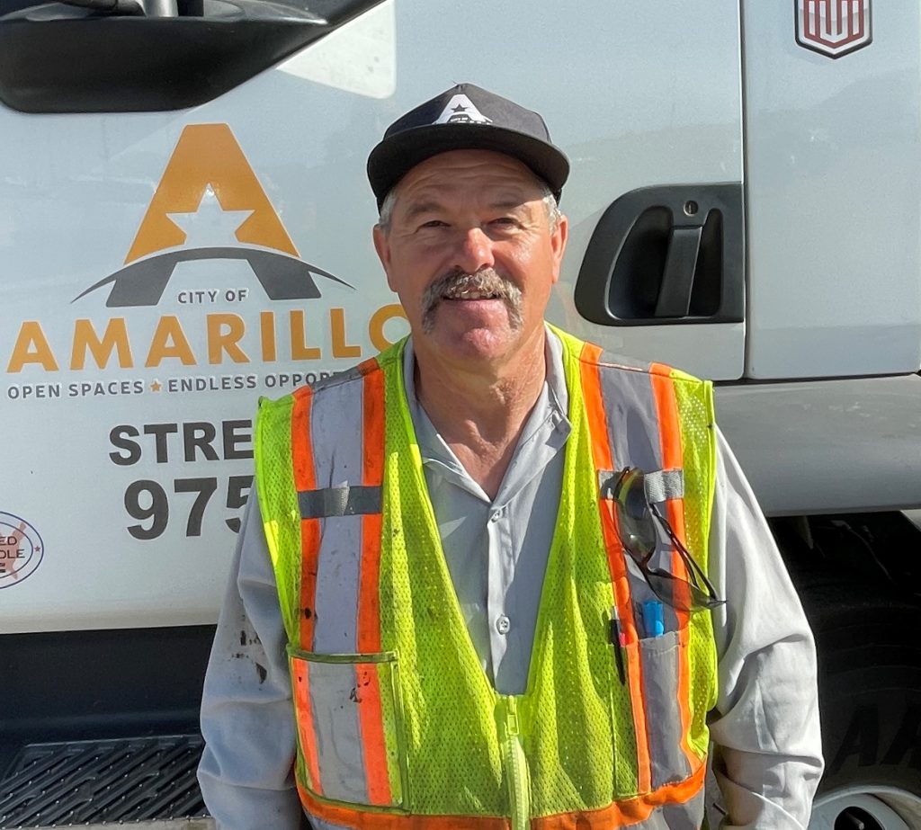 Paul Lundegreen, a City of Amarillo street department worker, stands smiling in front of a city service truck, wearing a high-visibility vest and a black Amarillo hat.