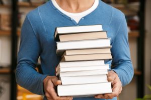 A man wearing a blue sweater holding a stack of various books