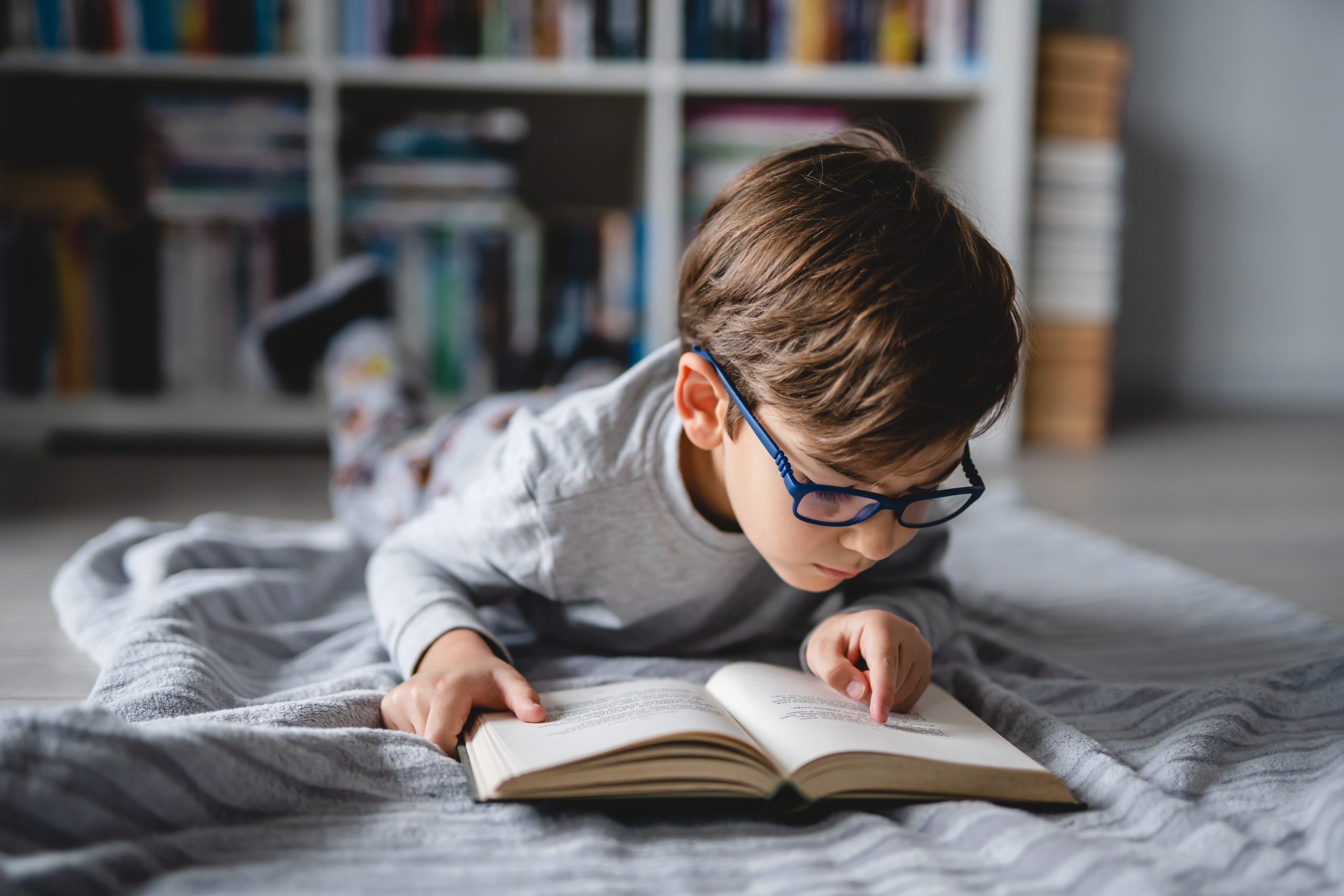 A young boy with glasses lying on the floor in his room reading a book