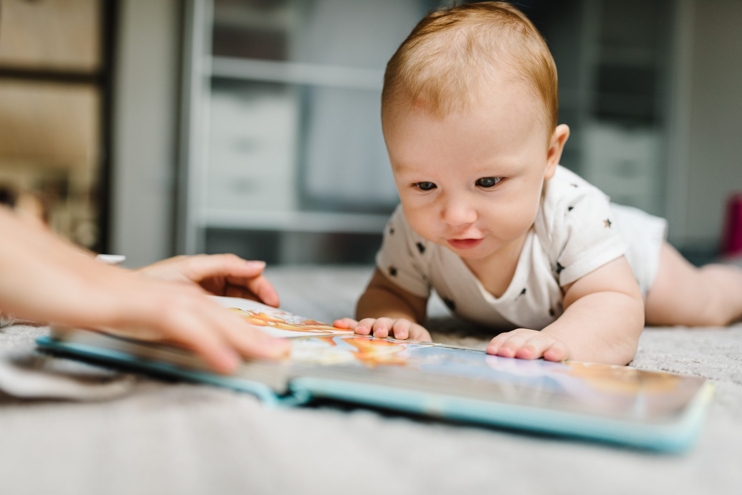 A young baby laying on their tummy reaching for an open children's book