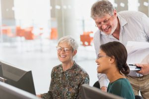 Two women smiling and working on a computer while an older man leans over to help