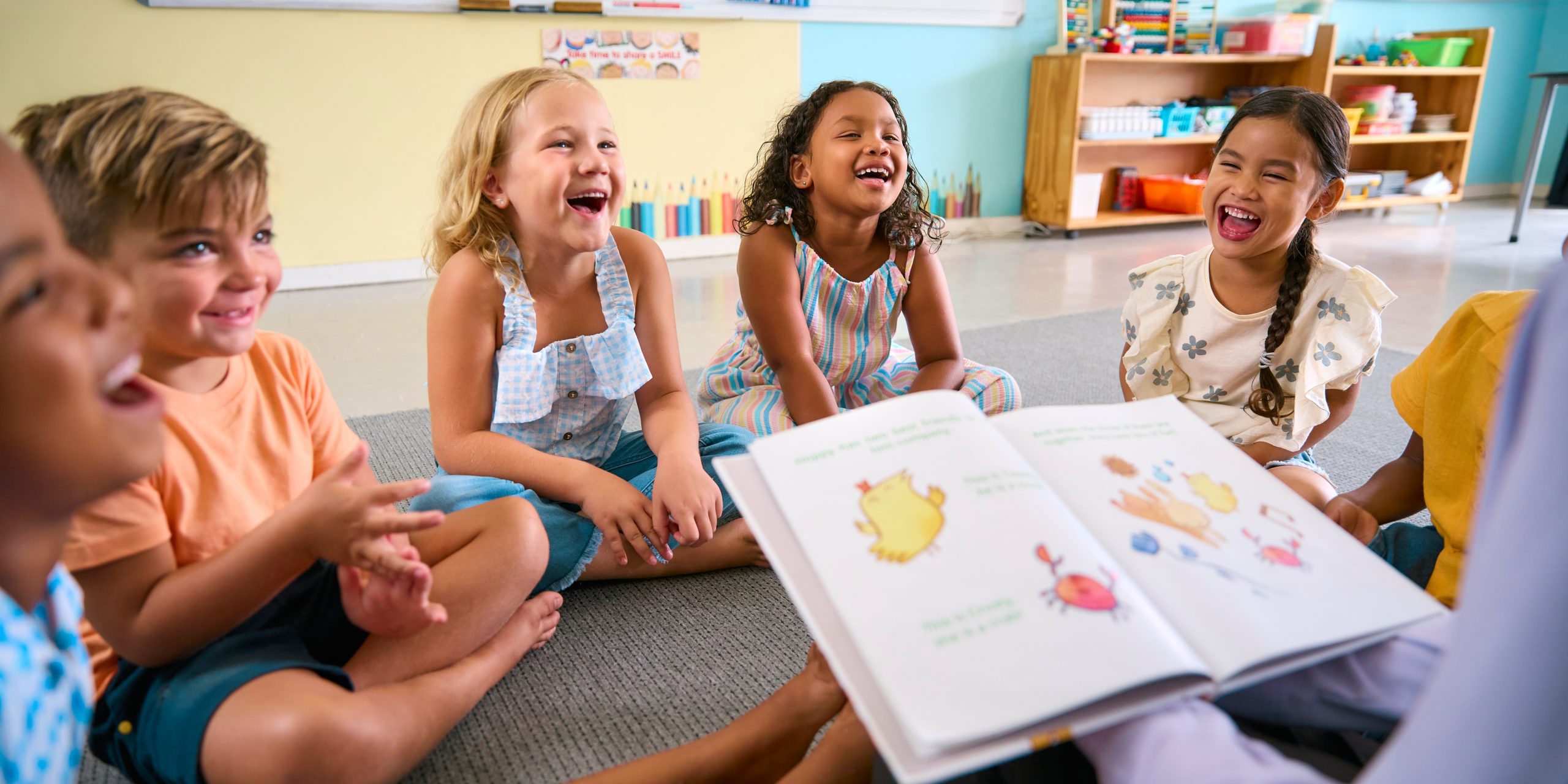 A group of children sitting on the ground laughing while someone reads them a book