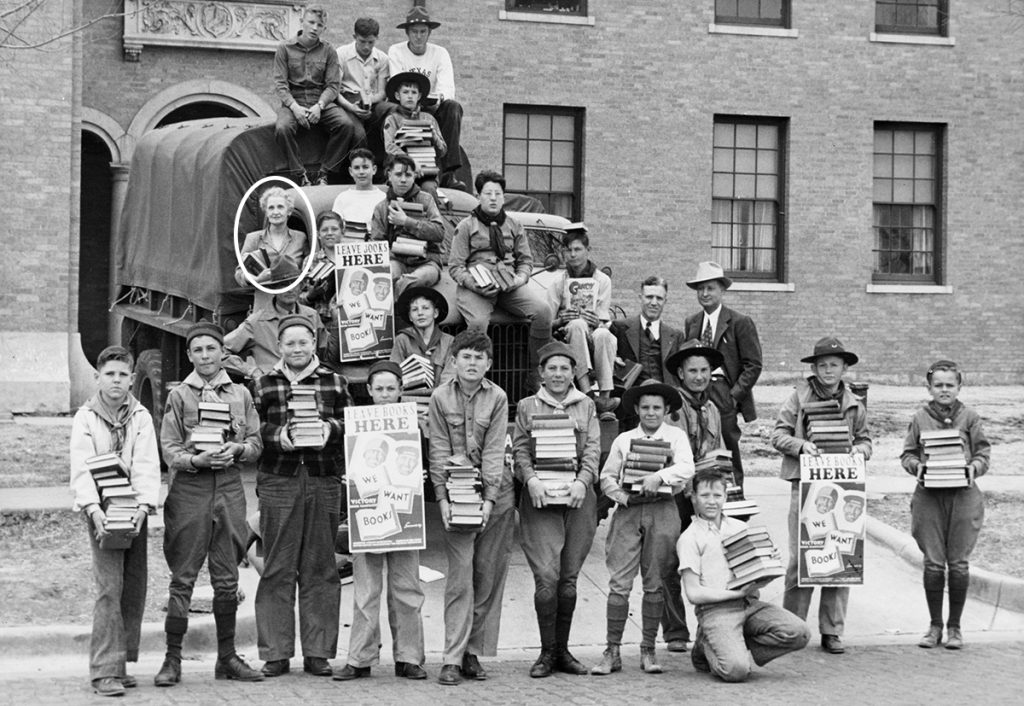 A vintage photo of Ruth Delzell and a group of boy scouts holding books and flyers reading "Leave Books Here"