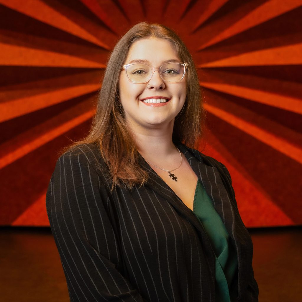 Headshot of Civic Center's Marketing Administrator Ashley Jones wearing glasses, a black pin-striped blazer, and a green shirt
