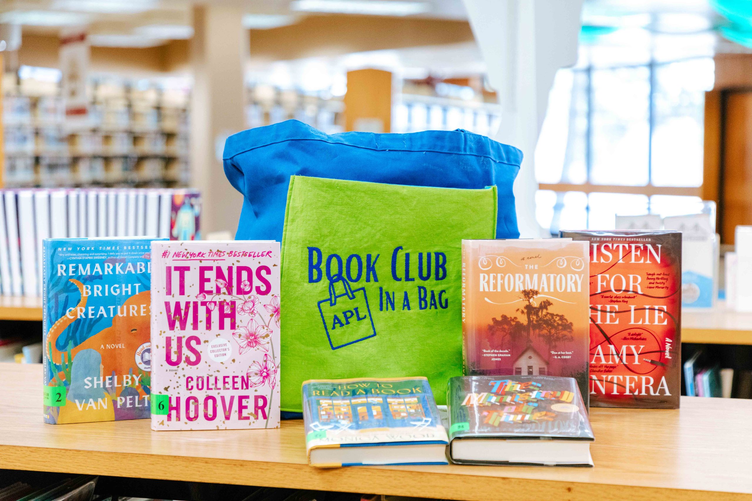 A blue tote bag with a green pocket in the front that reads "APL Book Club in a Bag" with six different books around the bag