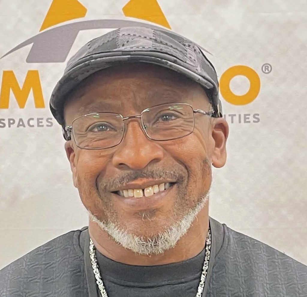 A headshot of Antonio Graves wearing wire glasses, a grey plaid hat, black shirt, and silver chained necklace