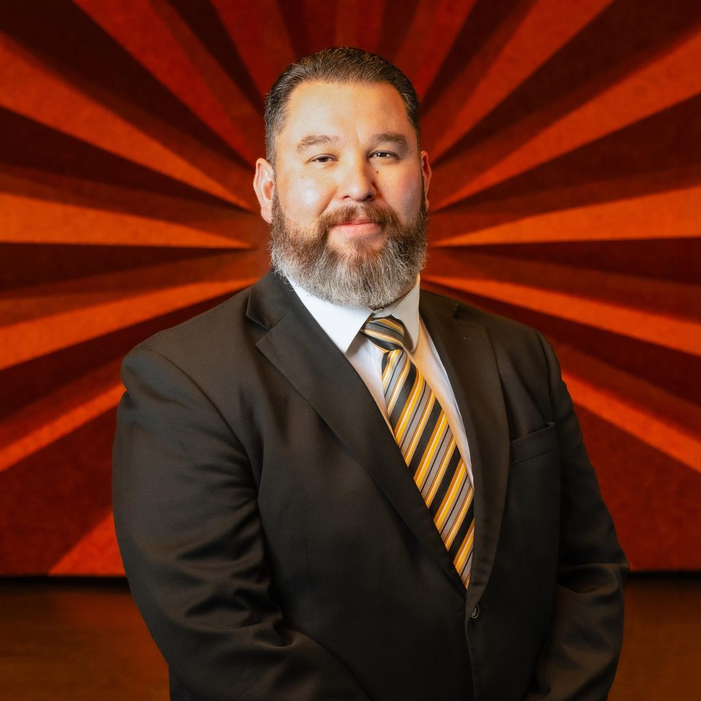 Headshot of Civic Center's Operations Manager David Balderaz wearing a black blazer and yellow striped tie