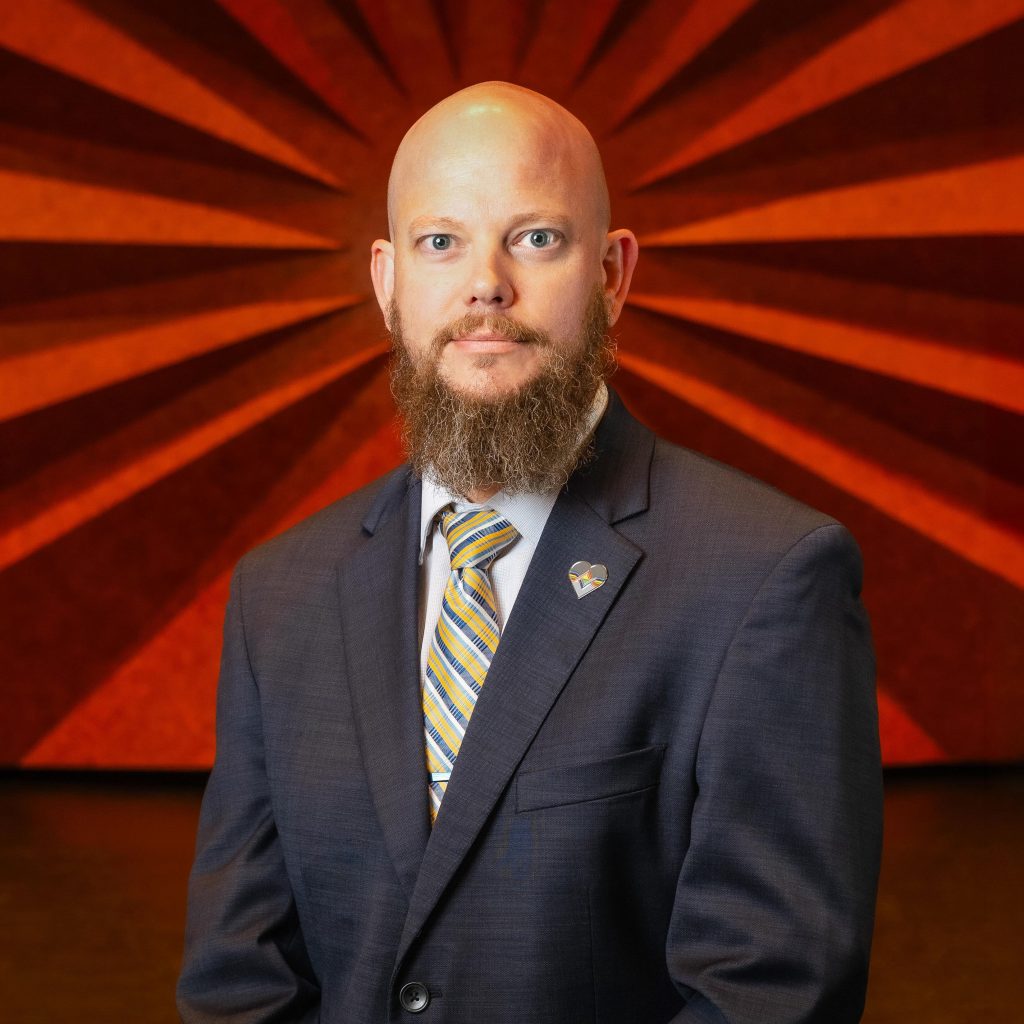 Headshot of Civic Center's Production Manager Derek Hisek wearing a black blazer and yellow striped tie