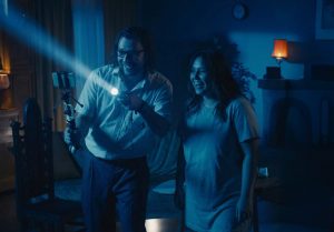 A man and woman in a dark room with a flashlight, smiling as they look out into the room for ghosts