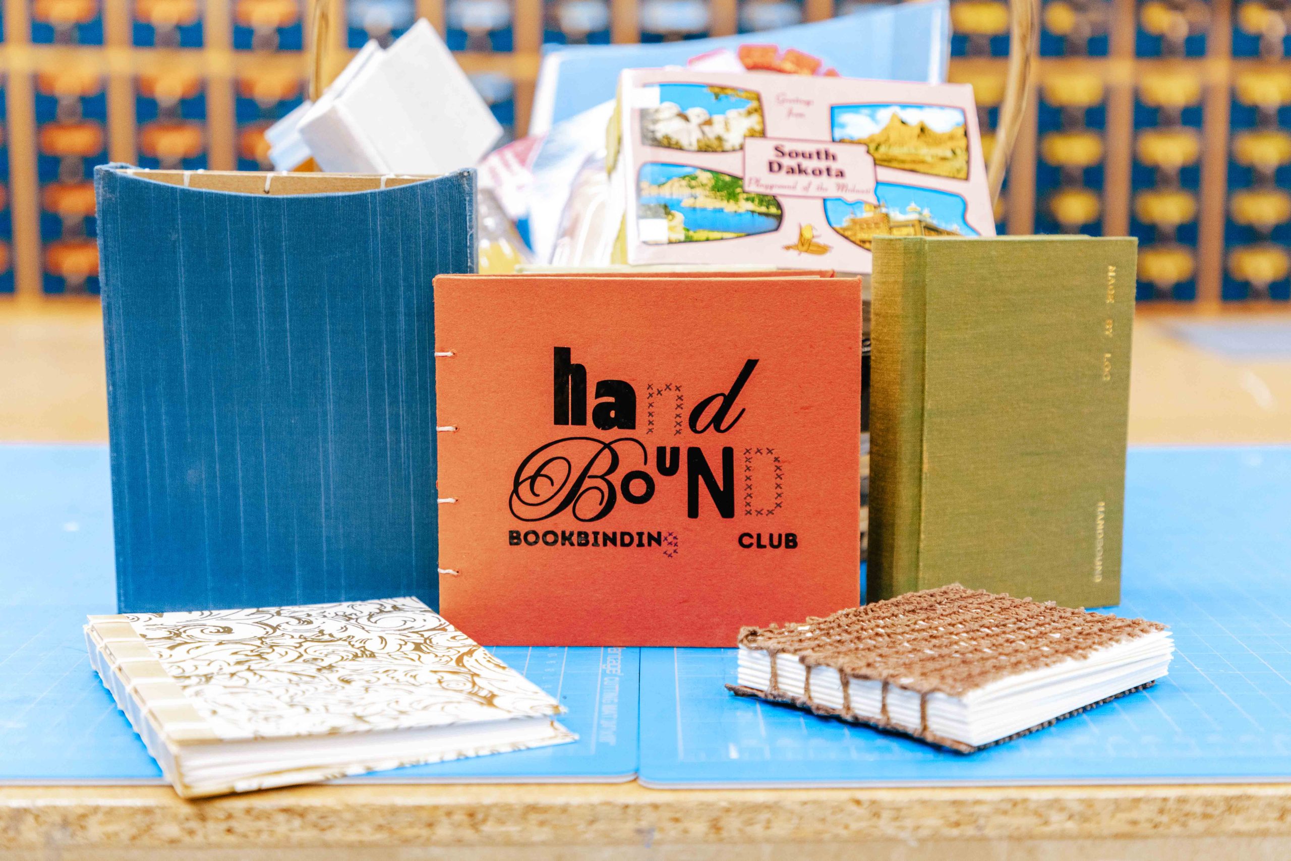 A group on hand-bound books in a variety of sizes and colors with the center book reading "Handbound Bookbinding Club"