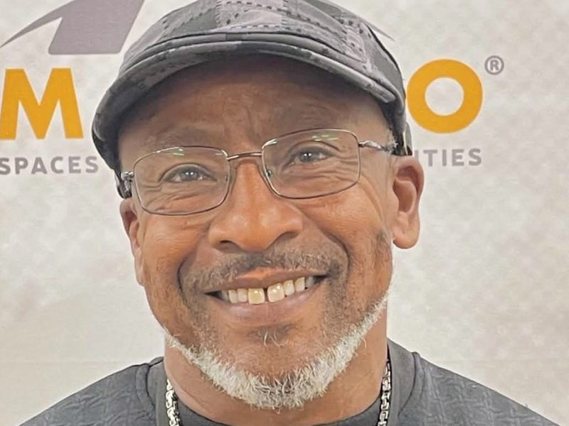 A headshot of Antonio Graves wearing wire glasses, a grey plaid hat, black shirt, and silver chained necklace